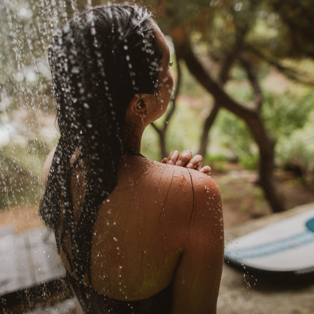 Woman with braids rinsing off in an outdoor shower, a surfboard beside her