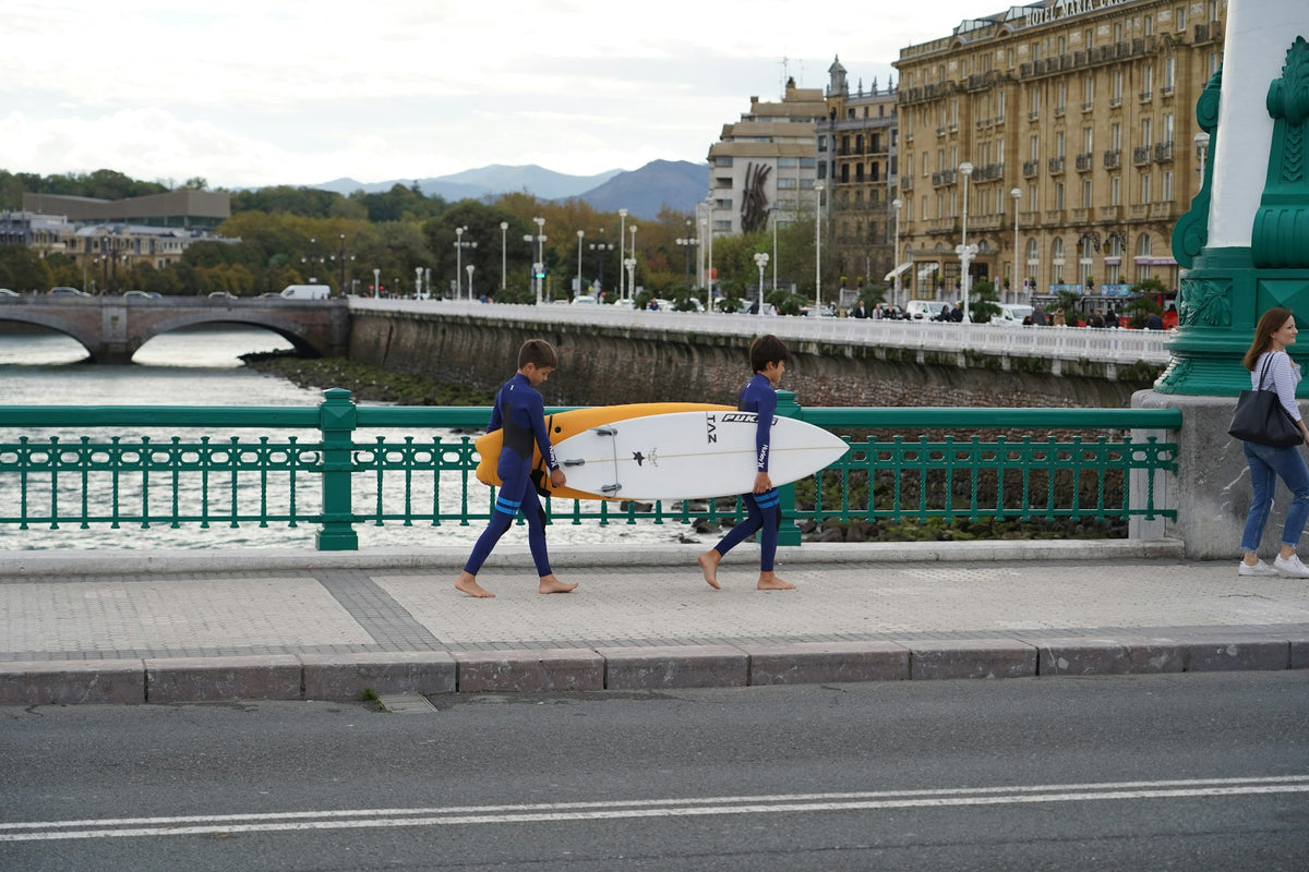 Two people walking along a seafront street in San Sebastián, one carrying a surfboard